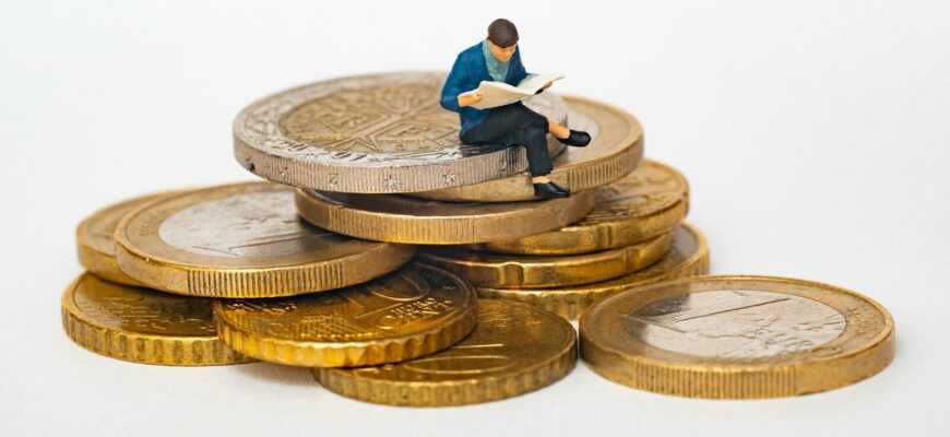 Miniature person sitting on stack of coins reading newspaper