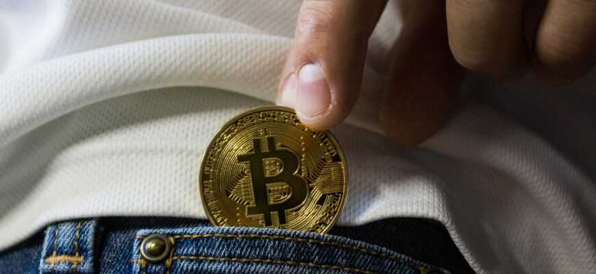 Close-up of hand putting Bitcoin in jeans pocket symbolizing cryptocurrency savings.